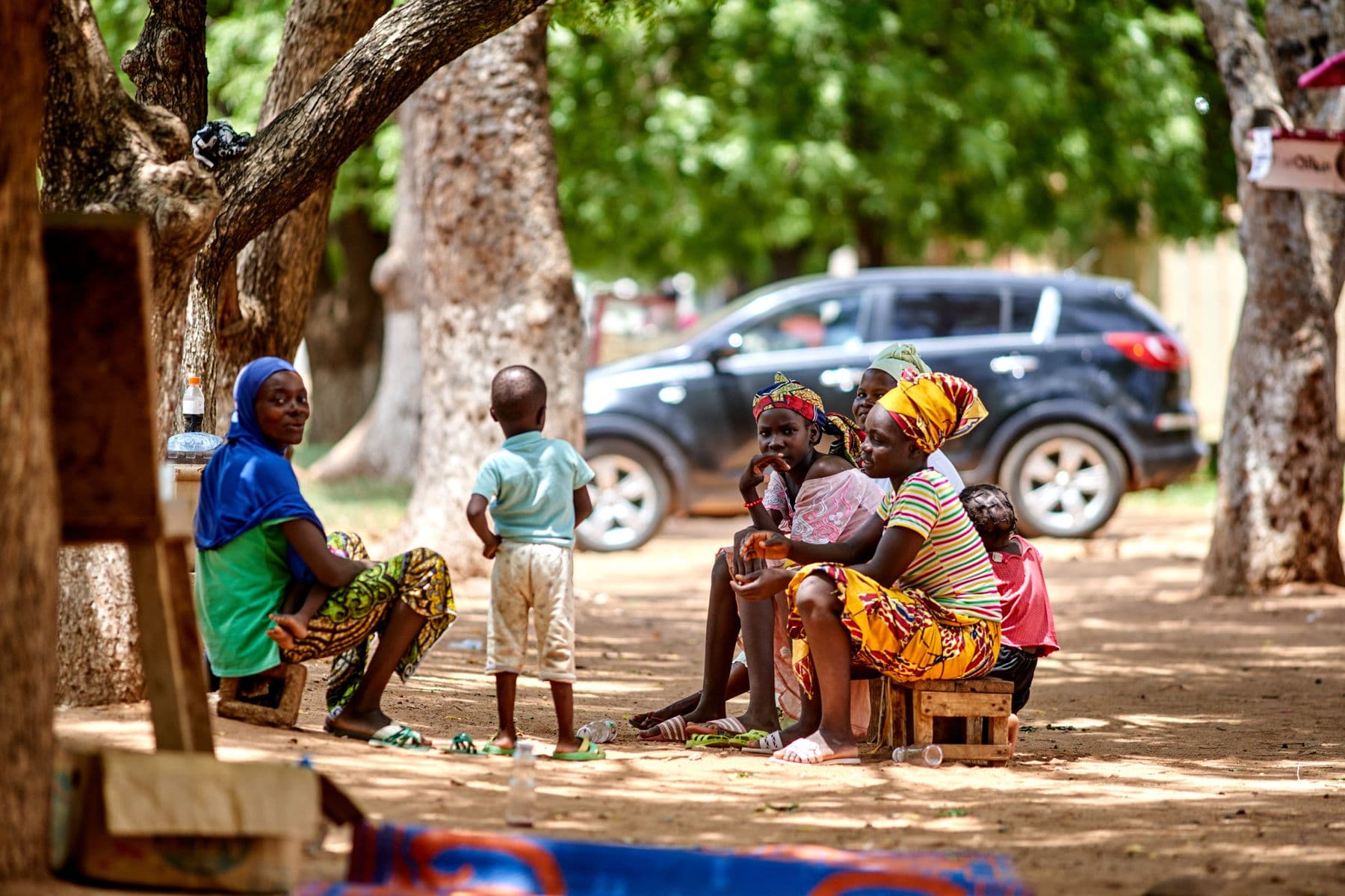 Women and children sit together under trees in a shaded community gathering.
